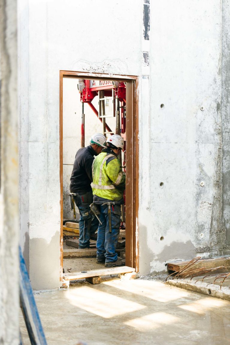 Deux ouvriers du bâtiment portant des casques EOVEST et des gilets de sécurité se tiennent devant une porte ouverte à l'intérieur d'un bâtiment partiellement construit. Ils travaillent avec des échafaudages et d'autres équipements de construction. Les murs environnants sont en béton brut et des planches de bois reposent sur le sol.