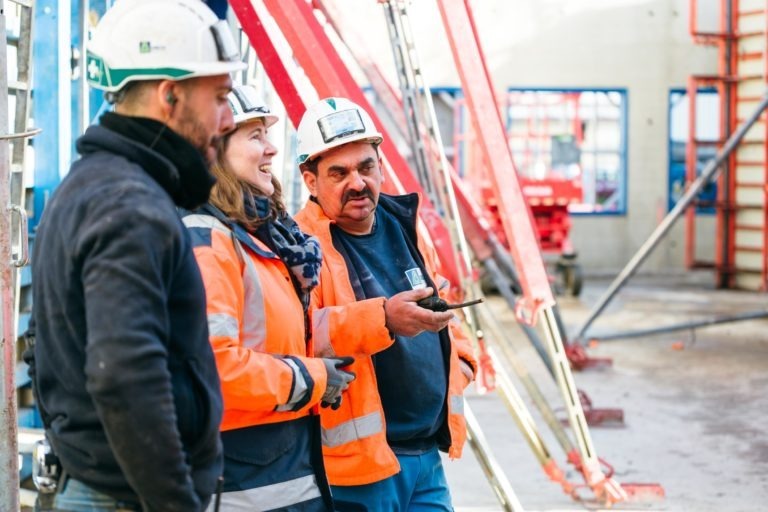 Trois ouvriers du bâtiment portant des gilets et des casques de haute visibilité, marqués du logo EOVEST, discutent sur un chantier. L'ouvrier de droite tient un téléphone et un échafaudage rouge est visible en arrière-plan. Tous les trois semblent engagés et concentrés sur leur conversation.