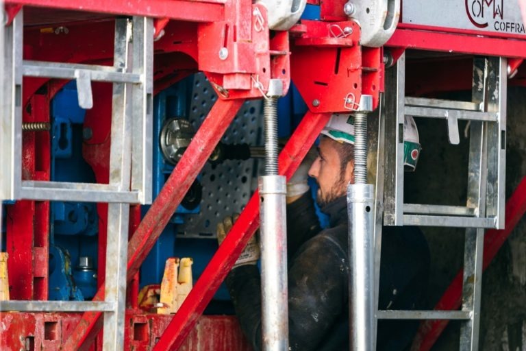 Un ouvrier du bâtiment portant un casque de sécurité et une veste de haute visibilité conduit une grande machine industrielle rouge et bleue avec la marque « EOVEST ». Le travailleur se concentre sur les commandes et est entouré de poutres métalliques et de composants structurels, garantissant que chaque détail répond aux normes de sécurité strictes.