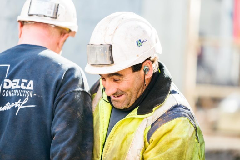 Deux ouvriers du bâtiment portant des casques blancs et des équipements de protection sur un chantier. L'ouvrier au premier plan, portant une veste jaune EOVEST et des protections auditives, sourit et regarde vers le bas. L'ouvrier à l'arrière-plan porte une veste noire avec l'inscription « Delta Construction » dans le dos.