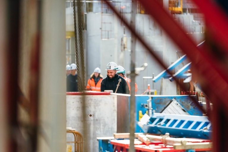 Un chantier de construction avec plusieurs ouvriers portant des casques de sécurité et des vêtements de haute visibilité. L'attention est portée sur un ouvrier portant un casque et une barbe blancs, debout à côté d'une machine industrielle EOVEST bleue et rouge. L'arrière-plan montre des équipements, des éléments structurels et d'autres ouvriers flous au loin.