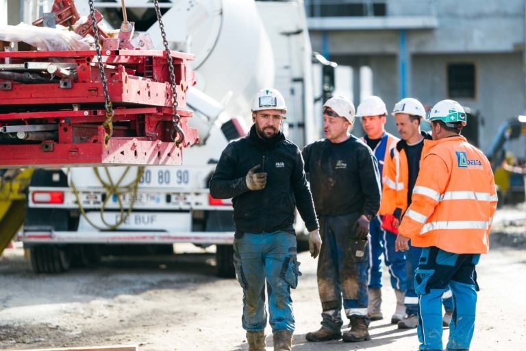 Un groupe de cinq ouvriers du bâtiment portant des équipements de sécurité et des casques EOVEST se tient sur un chantier de construction. Un ouvrier portant une veste noire et un pantalon bleu marche vers la caméra en tenant une boisson. D'autres, portant des gilets orange et bleus, observent une grue rouge qui soulève des matériaux. Un camion-citerne est visible en arrière-plan.