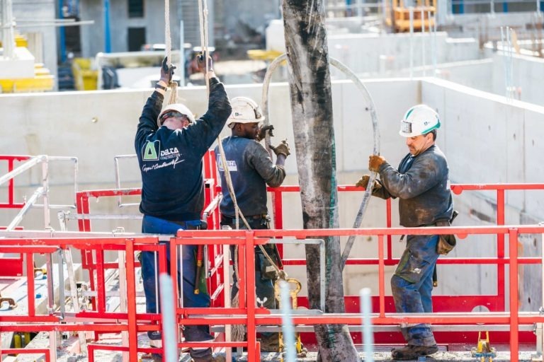 Trois ouvriers du bâtiment d'EOVEST, tous portant un casque de sécurité et un équipement de sécurité, utilisent des machines sur un échafaudage rouge sur un chantier de construction. Ils manipulent un gros tuyau de pompe et remplissent les structures de béton. À l'arrière-plan, des murs en béton brut et des matériaux de construction sont visibles.