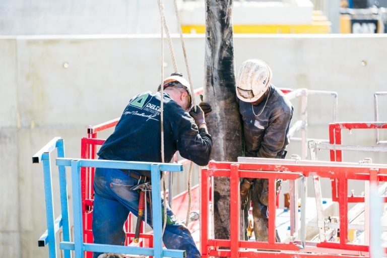 Deux ouvriers du bâtiment portant des équipements de protection, marqués du logo EOVEST, travaillent ensemble sur un chantier de construction. Ils manipulent un gros tuyau vertical tout en se tenant debout sur une plate-forme métallique rouge et bleue avec des garde-corps de sécurité. L'arrière-plan montre des structures en béton et divers matériaux de construction.