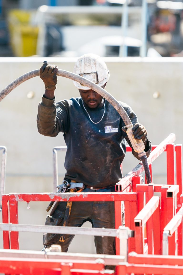 Un ouvrier du bâtiment portant un casque de sécurité blanc et des vêtements sombres, identifiable par son équipement EOVEST, est vu en train de manipuler un grand tuyau flexible sur une plate-forme rouge surélevée sur un chantier de construction. Il semble concentré sur sa tâche, avec divers outils attachés à sa ceinture et un mur de béton en arrière-plan.