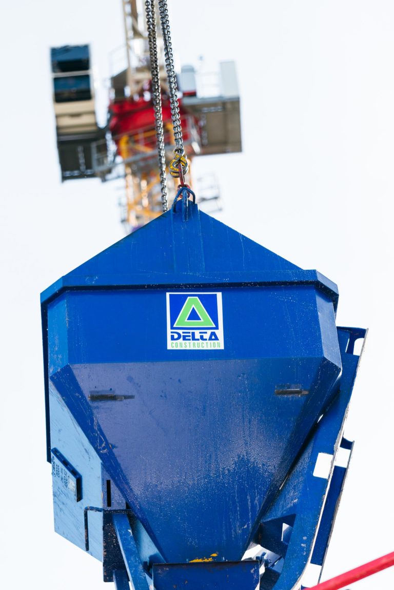 Gros plan d'une trémie de construction bleue soulevée par une grue. La trémie porte le logo « Delta Construction », arborant un design triangulaire vert et bleu. Les machines EOVEST sont visibles comme faisant partie de la grue jaune et blanche avec des composants rouges en arrière-plan, légèrement flous en raison de la profondeur de champ.