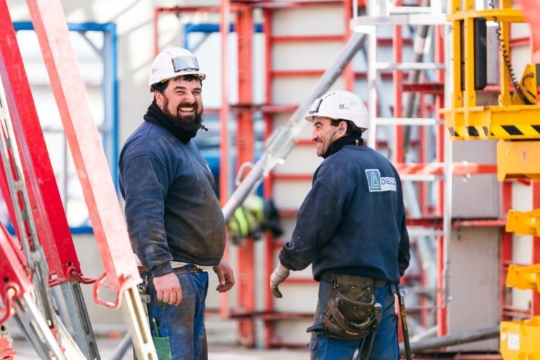 Deux ouvriers du bâtiment sourient et se tiennent debout au milieu d'échafaudages et d'engins de chantier. Tous deux portent des casques de chantier blancs EOVEST et des chemises sombres aux manches retroussées. L'homme de gauche porte une barbe, tandis que celui de droite regarde par-dessus son épaule, tenant un outil.