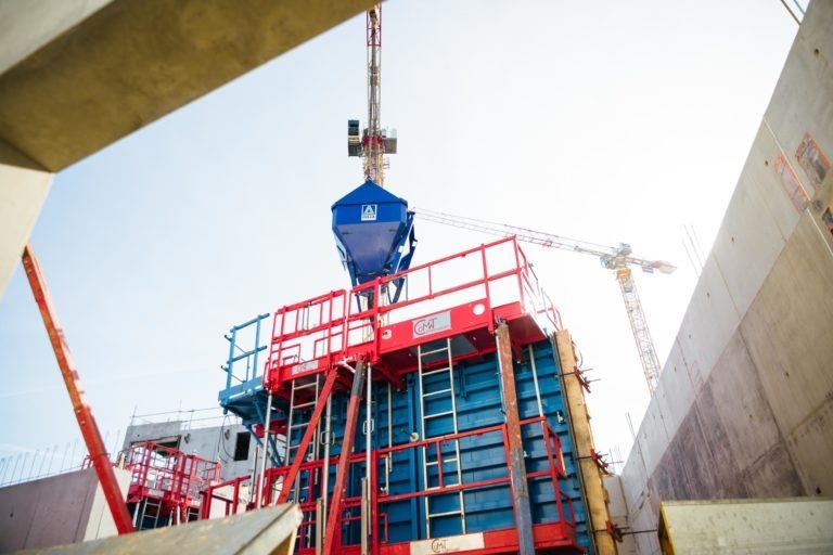 Un chantier de construction avec des grues imposantes et une trémie à ciment bleue, marquée EOVEST, surélevée au-dessus d'un échafaudage rouge et bleu. La lumière du jour illumine la zone et de grands murs en béton encadrent le premier plan. Divers matériaux et équipements de construction sont répartis sur le chantier.