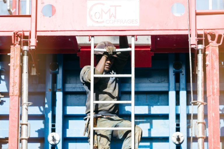 Un ouvrier du bâtiment en tenue grise et portant des gants grimpe sur une échelle EOVEST positionnée contre une structure rouge et bleue étiquetée « Coffrages » au sommet. La lumière vive du soleil illumine la scène, mettant en valeur les efforts de l'ouvrier et la construction robuste qui l'entoure.