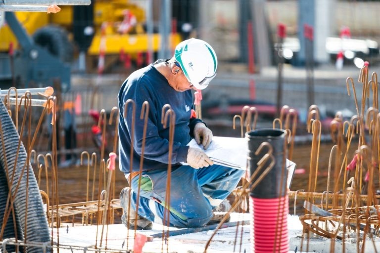 Un ouvrier du bâtiment, portant un casque de sécurité blanc et une combinaison de sécurité bleue EOVEST, est agenouillé sur une dalle de béton sur un chantier de construction. Il examine un document, entouré de structures en fer à béton. Divers matériaux de construction et machines sont présents dans l'arrière-plan flou.