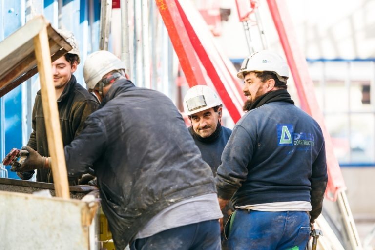 Quatre ouvriers du bâtiment portant des casques de sécurité et des vestes EOVEST sont rassemblés autour d'un engin de chantier sur un chantier. Ils semblent être en train de discuter ou d'effectuer une tâche. Des échafaudages et des matériaux de construction sont visibles en arrière-plan. La veste d'un ouvrier porte le texte « Delfio Construction ».