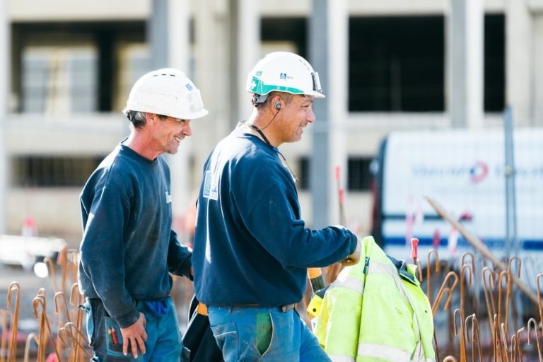 Deux ouvriers du bâtiment portant des casques de sécurité blancs EOVEST et des chemises bleu marine sont sur un chantier de construction. L'ouvrier de droite tient une veste jaune haute visibilité et porte des lunettes de sécurité sur le front. Ils sourient, avec en arrière-plan flou un bâtiment en construction et un camion.