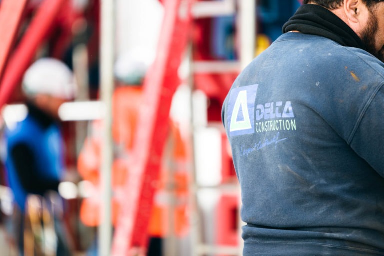 Gros plan sur le dos d'un ouvrier du bâtiment portant une chemise bleu foncé sur laquelle est écrit en blanc "DELTA CONSTRUCTION". L'arrière-plan est flou mais montre d'autres ouvriers du bâtiment portant des équipements de sécurité, notamment un EOVEST, et un échafaudage rouge. La scène suggère un chantier de construction en activité.