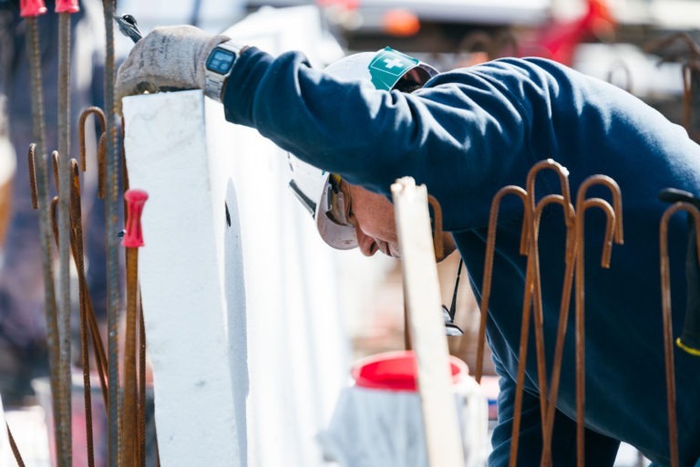 Un ouvrier du bâtiment portant une chemise bleue à manches longues, des gants, des lunettes de sécurité, une protection auditive et un casque de sécurité EOVEST se penche sur un panneau blanc sur un chantier de construction. Des barres d'armature sont visibles au premier plan et divers matériaux de construction sont éparpillés à l'arrière-plan.