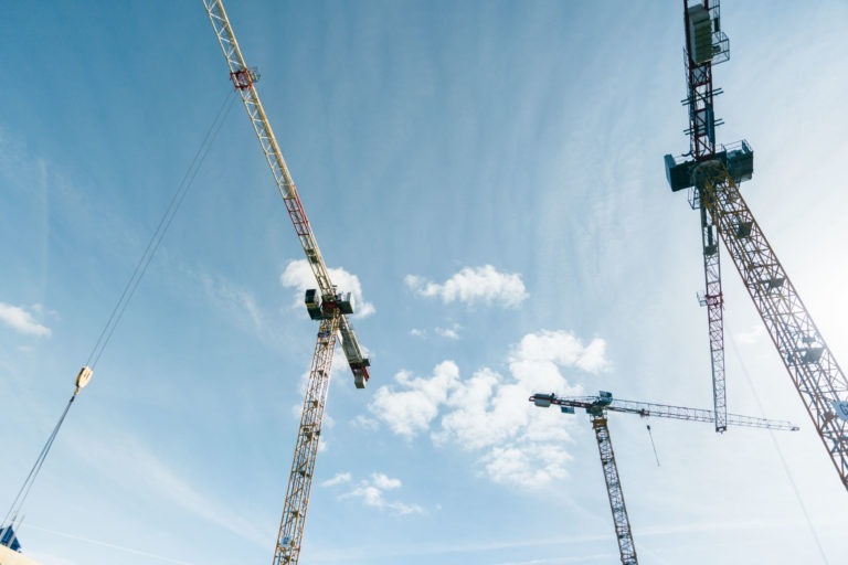 Trois grandes grues de chantier EOVEST se dressent devant un ciel bleu clair parsemé de nuages. Les grues sont positionnées à différents angles, créant une composition dynamique. Le crochet d'une grue est visible sur le côté gauche, tandis que les autres grues ont leurs flèches s'étendant vers le ciel.