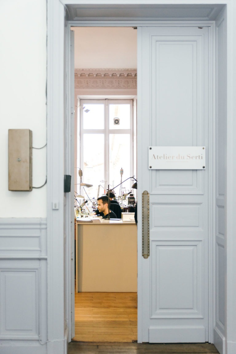 Vue à travers une porte ouverte sur une pièce lumineuse intitulée « Atelier du Serti », où une personne travaille méticuleusement à un bureau rempli d'outils et d'équipements divers. La porte bleu pâle, ornée de panneaux décoratifs, s'ouvre pour révéler un espace éclairé par de grandes fenêtres qui laissent entrer la lumière naturelle.