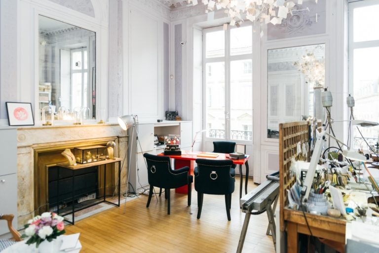 Une pièce chic et lumineuse avec de grandes fenêtres, avec deux chaises noires et une table rouge devant une cheminée décorative. La pièce, qui rappelle un Atelier du Serti, est remplie d'un décor éclectique comprenant un lustre élégant, des œuvres d'art, un miroir pleine longueur et divers objets sur des étagères et un bureau. Les planchers en bois clair ajoutent de la chaleur.