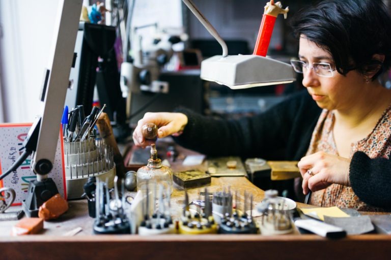 Une personne portant des lunettes travaille méticuleusement sur l'établi encombré du bijoutier de l'Atelier du Serti. L'espace de travail est éclairé par une lampe brillante et divers outils et équipements, dont un étau, des pinces et des tiges métalliques, sont disposés. La personne se concentre sur un petit objet dans sa main, avec précision et soin.