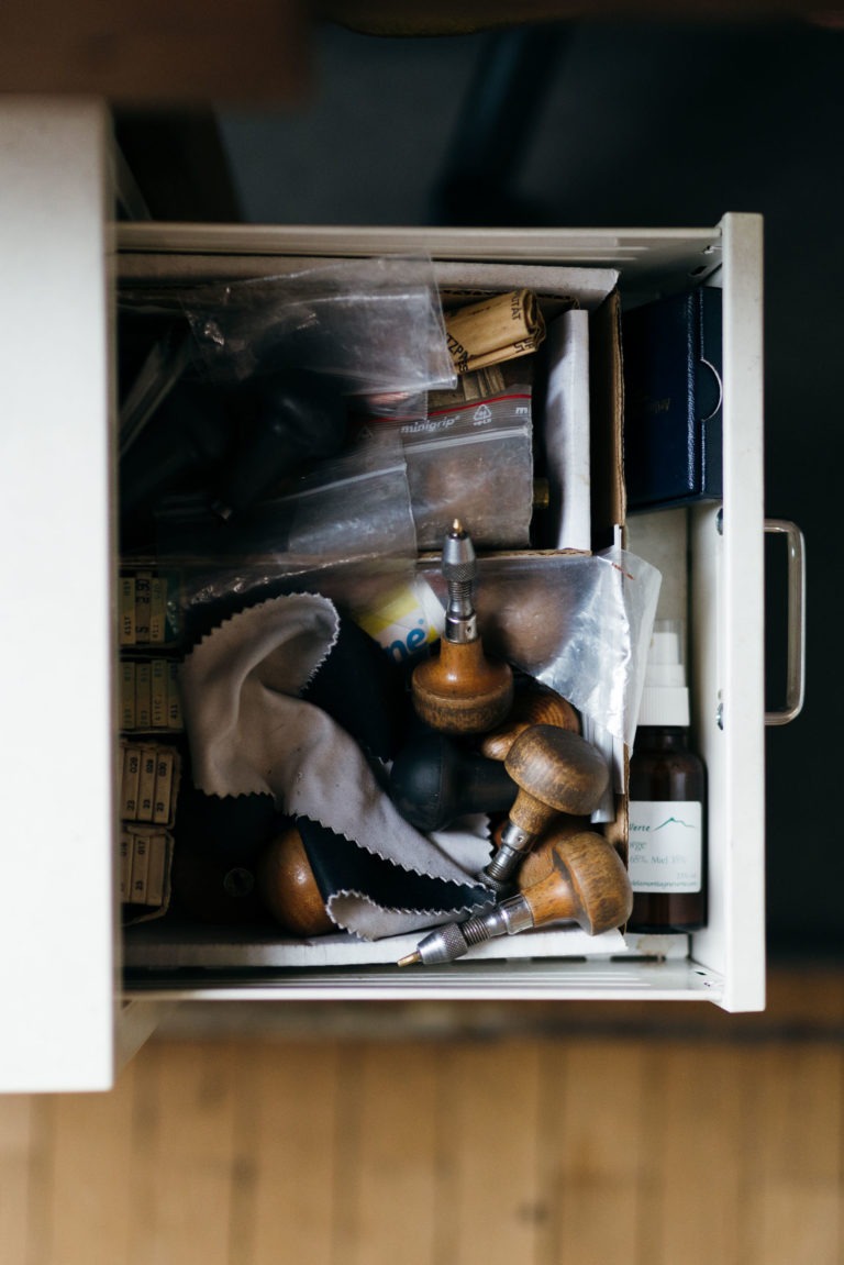 Un tiroir ouvert rempli d'outils et d'objets divers, caractéristique d'un espace de travail de l'Atelier du Serti. Il contient des outils en bois portatifs, une petite bouteille de liquide avec compte-gouttes, des sacs en plastique et un chiffon noir et blanc plié. Le tiroir blanc avec une poignée métallique est situé à l'intérieur sur un plancher en bois.