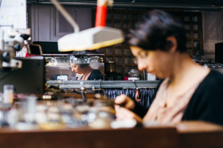 Une femme aux cheveux courts et foncés se concentre sur une tâche dans un Atelier du Serti rempli d'outils et d'équipements. Elle est au premier plan, légèrement floue, tandis que son reflet est clairement visible dans un miroir à l'arrière-plan, capturant sa concentration sur son travail.