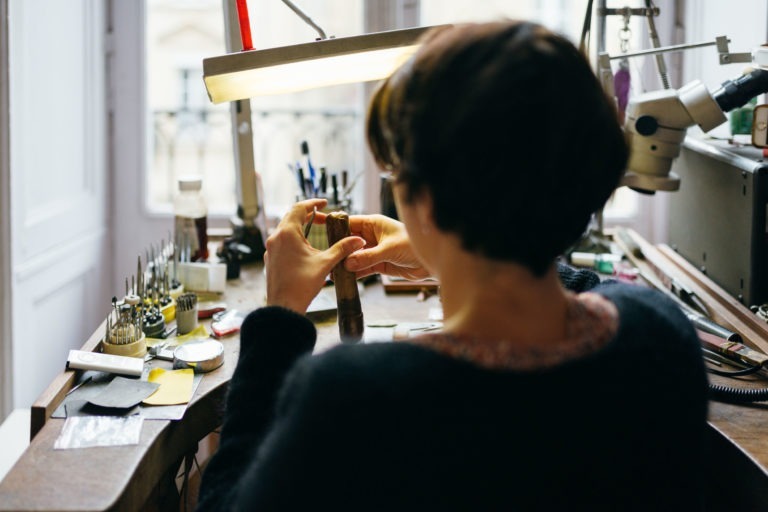 Une personne aux cheveux courts et foncés travaille à un bureau encombré sous une lampe lumineuse. Elle tient un objet cylindrique de couleur laiton et utilise des outils de la collection de l'Atelier du Serti. L'espace de travail comprend divers petits objets, des papiers et des récipients. L'arrière-plan montre des fenêtres floues et de la lumière qui entre.