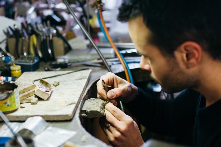 Un homme travaille avec attention sur un bijou sur un établi encombré rempli d'outils et de matériaux divers. Dans son Atelier du Serti, il tient délicatement le bijou dans une main et utilise un petit outil dans l'autre. L'arrière-plan est flou, concentrant l'attention sur son savoir-faire précis.