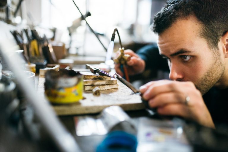 Un homme, concentré et déterminé, travaille sur une pièce de métal dans l'Atelier du Serti. Il utilise un fer à souder à deux mains, se concentrant intensément sur la tâche. L'espace de travail est encombré d'outils, dont une petite boîte de conserve, des pièces de métal et divers équipements, suggérant un projet détaillé et complexe.