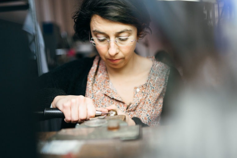 Une personne aux cheveux courts et foncés et portant des lunettes est concentrée sur le travail avec un outil, probablement dans un atelier de l'Atelier du Serti. Elle porte une blouse à motifs avec un cardigan. Ses mains tiennent une pièce sur laquelle elle travaille. L'arrière-plan est flou, gardant l'attention sur sa concentration et son activité.