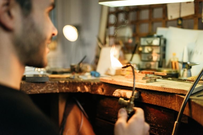 Une personne tenant une petite torche avec une flamme vive travaille sur un établi en bois encombré dans l'Atelier du Serti. L'espace de travail est rempli de divers outils et matériaux, tandis qu'une petite lampe fournit un éclairage supplémentaire. L'arrière-plan comprend d'autres outils et matériaux, suggérant un décor artisanal.