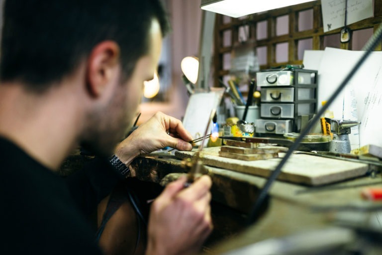 Un homme assis à un poste de travail bien éclairé dans l'Atelier du Serti tient des outils, se concentrant sur un petit objet. Le bureau est encombré de divers instruments, de papiers et de petits tiroirs. L'arrière-plan comprend des décorations épurées et un organiseur en grille de bois. La scène suggère un travail artisanal méticuleux et détaillé.