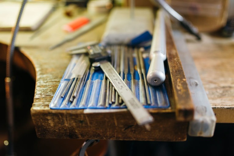 Un établi avec divers outils dispersés sur un tapis à outils bleu, notamment des pieds à coulisse, des forets et un manche en argent dans un décor Atelier du Serti. La surface de l'établi semble bien utilisée avec le grain du bois et d'autres outils flous en arrière-plan.