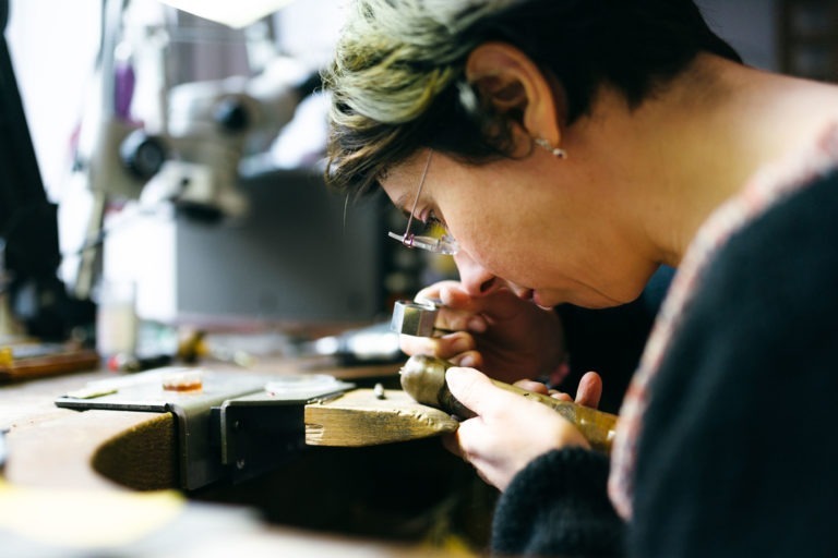 Une personne aux cheveux courts et portant des lunettes se concentre sur la fabrication d'un bijou sur un établi de l'Atelier du Serti. Elle tient un petit marteau dans une main et une pièce de métal dans l'autre, entourée de divers outils. L'espace de travail est bien éclairé, avec un microscope et d'autres équipements en arrière-plan.