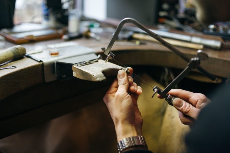 Gros plan sur les mains d'une personne qui coupe avec une scie sauteuse une pièce de bois posée sur un établi. L'Atelier du Serti est encombré de divers outils et objets. Les mains de la personne sont stables, concentrées sur la coupe précise. L'arrière-plan est légèrement flou, ce qui met en valeur l'activité de travail du bois.