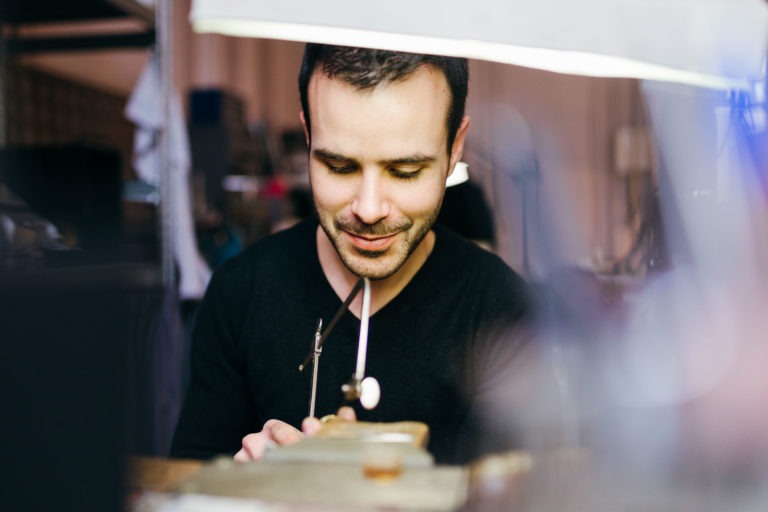 Une personne vêtue d'une chemise noire travaille sur une table encombrée de l'Atelier du Serti, remplie d'outils et de matériaux divers. Elle utilise une petite scie pour couper quelque chose et son expression faciale montre sa concentration. L'arrière-plan est flou, avec des objets indistincts et un éclairage doux créant une atmosphère calme.