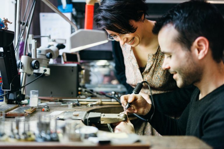 Deux personnes travaillent ensemble dans un atelier bien équipé. La personne à droite, un homme, utilise un outil portatif sur un bijou tandis que la femme à gauche regarde et sourit. L'espace de travail encombré est rempli d'outils et de matériaux divers, incarnant l'esprit du savoir-faire de l'Atelier du Serti.