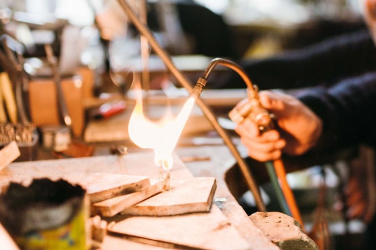 Une personne utilise un chalumeau à flamme visible pour travailler sur une pièce de métal dans l'atelier du Serti. L'espace de travail est encombré d'outils et de matériaux divers. L'arrière-plan est flou, suggérant un espace fréquenté et bien utilisé. La flamme intense du chalumeau est le point focal de l'image.