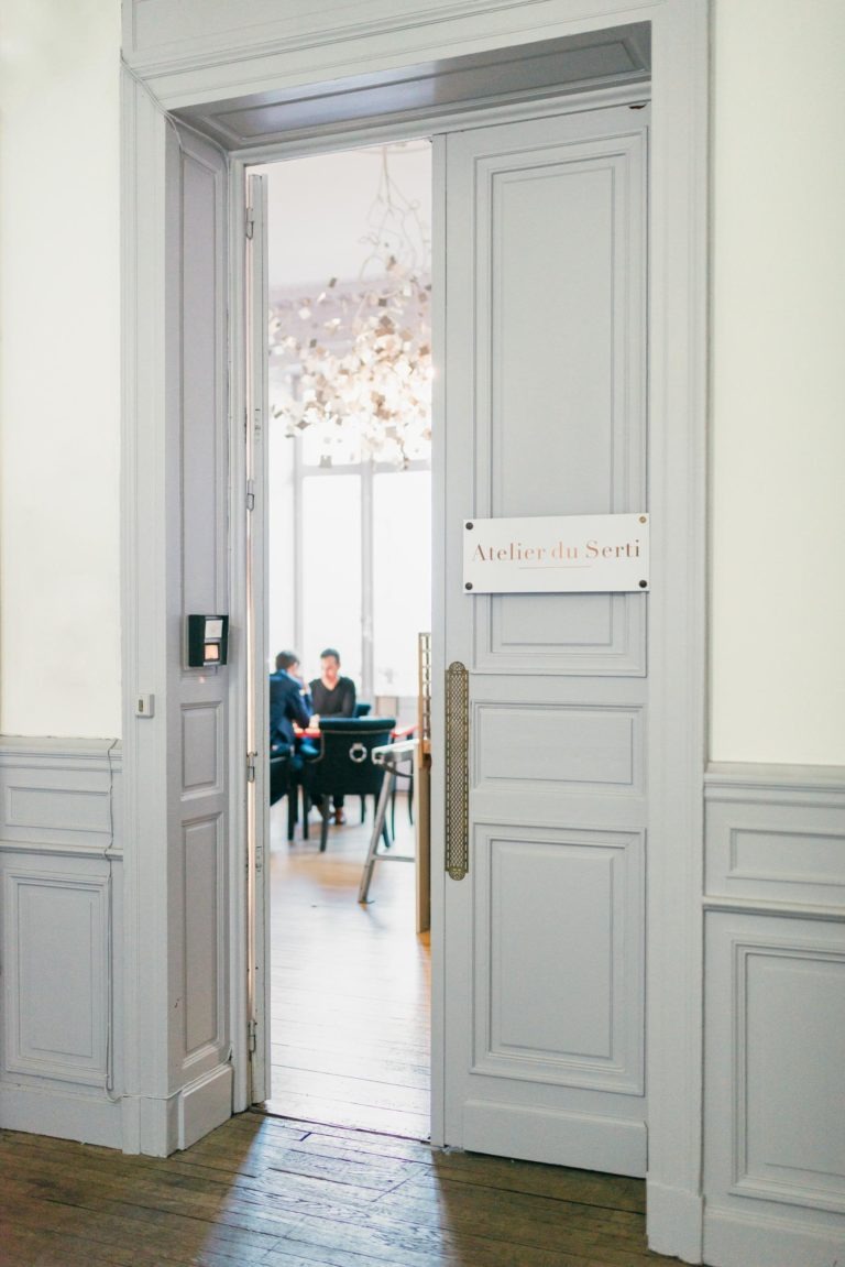 Une porte entrouverte mène à une pièce lumineuse intitulée « Atelier du Serti ». Deux personnes sont assises à une table en arrière-plan près d'une grande fenêtre. La pièce, aux murs gris clair et au parquet en bois, respire l'élégance avec un lustre orné de nombreux éléments suspendus au-dessus de la table.