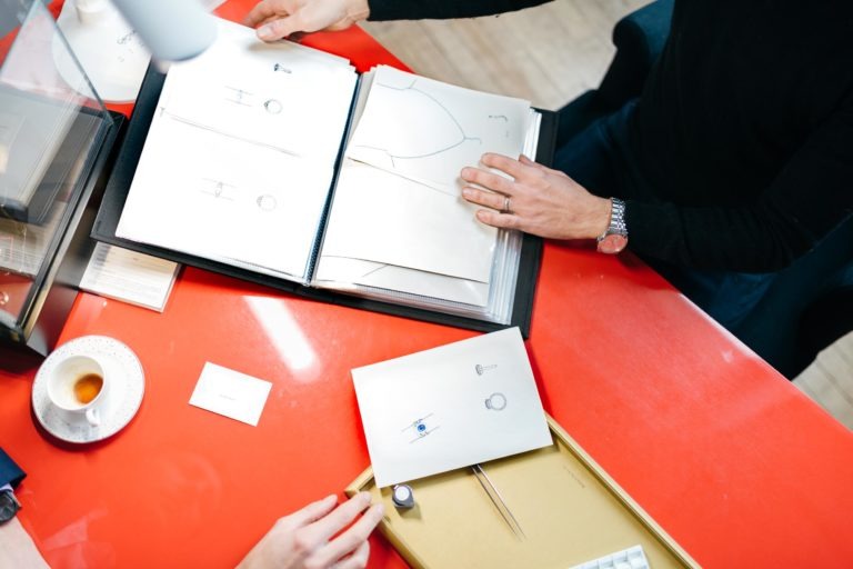 Deux personnes sont assises à une table rouge dans l'Atelier du Serti, examinant des croquis de conception dans un dossier. Une personne tient le dossier, tandis que l'autre fait un geste dans sa direction. Divers outils, une bague, une tasse à thé et une carte de visite sont posés sur la table, suggérant une séance de travail collaborative sur la conception de bijoux.