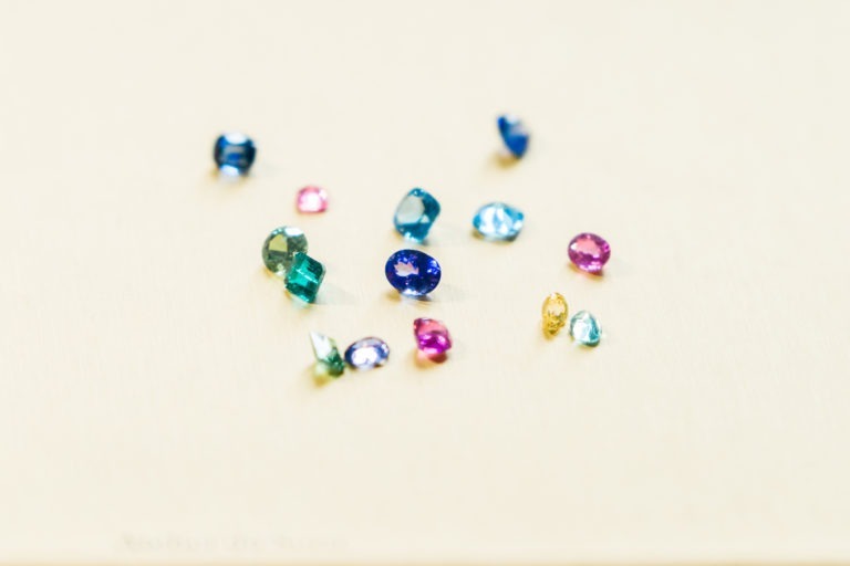 Une collection de petites pierres précieuses colorées sur une surface beige clair. Les pierres varient en forme et en couleur, notamment en bleu, vert, rose, jaune et violet. Chaque pierre est polie et possède une taille à facettes, mettant en valeur leur éclat et réfléchissant la lumière, un véritable chef-d'œuvre de l'Atelier du Serti.