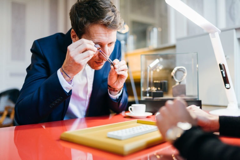 Un homme en costume bleu marine examine une montre à travers une loupe, assis à une table rouge ornée d’outils de l’Atelier du Serti. Sur la table se trouvent un plateau jaune, un clavier blanc, une tasse à café blanche et une soucoupe. La main d’une autre personne est visible au premier plan. À l’arrière-plan, il y a une vitrine en verre avec une autre montre à l’intérieur.