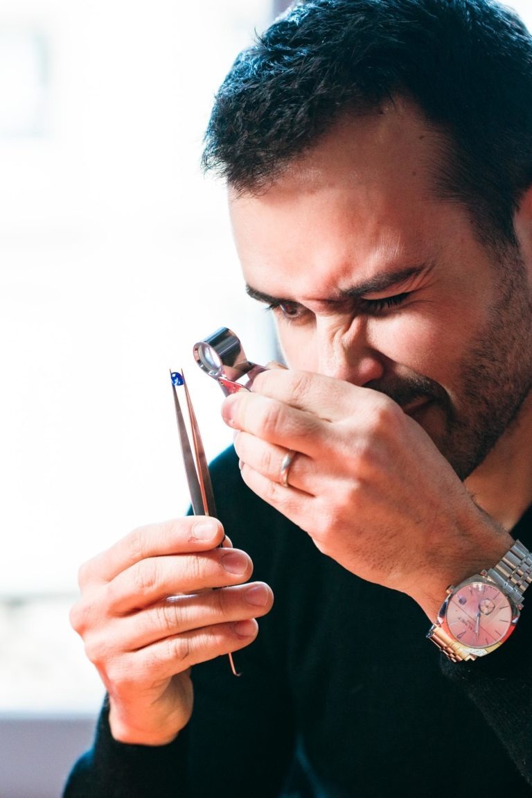 Un homme aux cheveux noirs courts examine une petite pierre précieuse bleue à l'aide d'une loupe de bijoutier tenue contre son œil. Il tient la pierre précieuse avec une pince à épiler dans une main, semblant concentré. Vêtu d'une chemise noire et d'une montre-bracelet en argent, il travaille probablement dans un Atelier du Serti, avec un arrière-plan flou derrière lui.