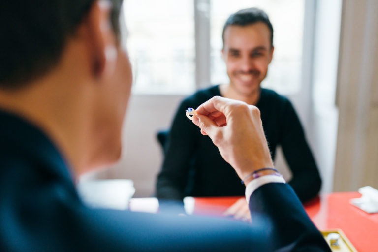 Gros plan de deux personnes, l'une vêtue d'un costume sombre, qui est floue au premier plan et qui tient un petit objet, peut-être une bague de l'Atelier du Serti. L'autre personne, nette à l'arrière-plan, porte une chemise noire et sourit, assise à une table rouge. Le décor semble être intérieur avec un éclairage tamisé.