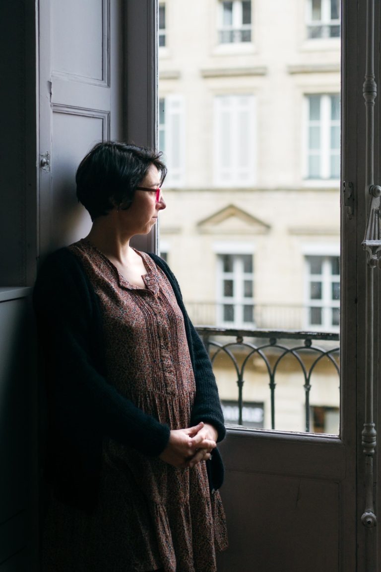 Une personne aux cheveux courts et foncés, portant des lunettes, une robe à motifs marron et un cardigan noir, se tient près d'une fenêtre donnant sur l'extérieur. Ils ont les mains jointes et s'appuient contre le mur. À l'arrière-plan, on peut voir un bâtiment de l'Atelier du Serti avec de nombreuses fenêtres et des balustrades de balcon.