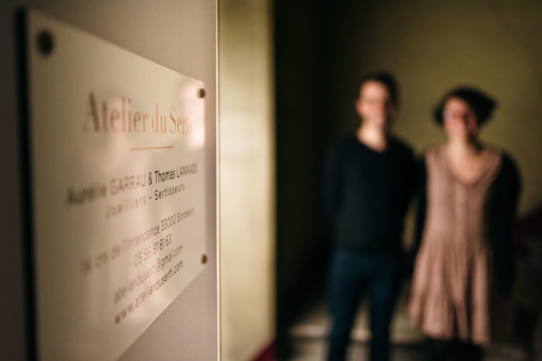 Une image floue de deux personnes debout dans un couloir faiblement éclairé. Au premier plan, on peut voir un panneau indiquant clairement « Atelier du Séra » avec des noms et des coordonnées en dessous, ce qui fait peut-être allusion à son autre nom « Atelier du Serti ». L'arrière-plan est flou, ce qui rend difficile de discerner les traits détaillés des individus.