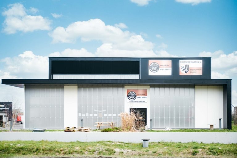 Un bâtiment rectangulaire moderne avec des enseignes pour une brasserie et un restaurant se dresse fièrement à Pessac. La façade est blanche avec une grande section vitrée au milieu. L'enseigne de la brasserie affiche un logo circulaire avec du texte, tandis que l'enseigne du restaurant indique « Brasserie Gasconha ». Des plantes en pot et des bancs bordent la façade sur l'herbe verte, sous un ciel bleu parsemé de nuages.