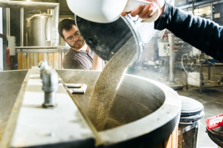 Un homme portant des lunettes verse un récipient de céréales dans une grande cuve de brassage industrielle à la Brasserie Gasconha de Pessac. La scène se déroule dans la brasserie avec divers équipements et tuyaux en acier inoxydable visibles en arrière-plan. De la vapeur s'élève de la cuve, créant une atmosphère visiblement chaleureuse.