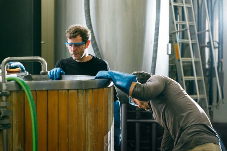 Deux hommes en tenue de protection, dont des gants bleus et des lunettes de sécurité, travaillent à la Brasserie Gasconha de Pessac. L'un inspecte une grande cuve en bois tandis que l'autre se penche dessus. Des équipements industriels, notamment des cuves métalliques et une échelle, sont visibles en arrière-plan.