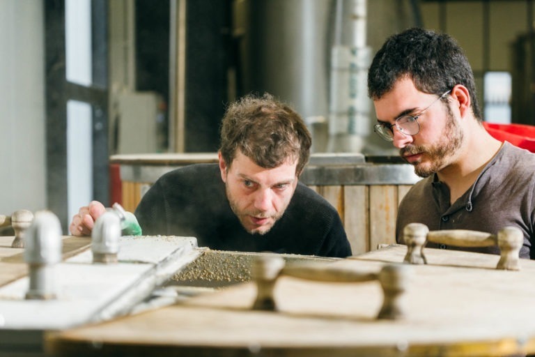 Deux hommes inspectent de près une machine de traitement des céréales dans un environnement industriel à la Brasserie Gasconha de Pessac. L'un verse de la poudre verte à partir d'un petit récipient, tandis que l'autre regarde attentivement. Tous deux portent des vêtements décontractés de couleur sombre et des lunettes. Divers équipements et machines sont visibles en arrière-plan.