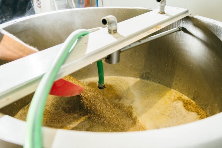 Gros plan sur une cuve de brassage pendant le processus de fabrication de la bière à la Brasserie Gasconha Pessac. Un tuyau vert et une palette métallique sont insérés dans la cuve où les grains sont mélangés à l'eau chaude. Le mélange apparaît mousseux, ce qui indique un brassage actif. De la vapeur s'élève de la cuve, créant un effet brumeux.