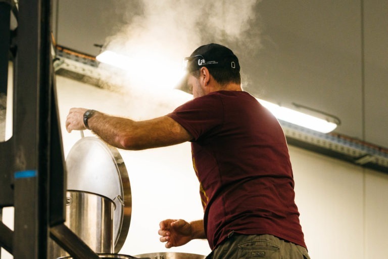 Un homme portant une casquette noire et un t-shirt marron soulève le couvercle d'une grande cuve en acier inoxydable, libérant de la vapeur dans l'air. À l'intérieur de la Brasserie Gasconha Pessac, il travaille sous un haut plafond avec des lumières fluorescentes. Une structure métallique noire est partiellement visible sur le côté gauche de l'image.